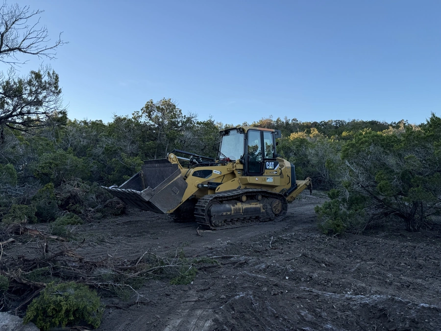 A yellow Caterpillar bulldozer with tracks sits in a wooded area, clearing debris and vegetation to prepare the site for construction.