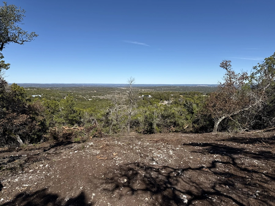 A scenic overlook on a dirt trail in a wooded, hilly area, with a panoramic view of the surrounding countryside stretching out in the distance under a bright blue sky.