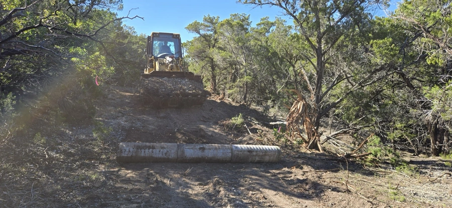 A large bulldozer clearing a path through a wooded area, with fallen branches and debris scattered around.