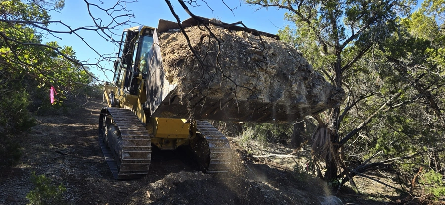 A heavy-duty construction excavator with a large bucket is clearing a path through dense vegetation and branches in a wooded area, kicking up dirt and debris as it moves forward.