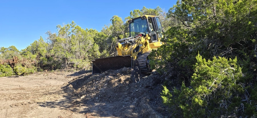 A Caterpillar bulldozer clearing a path through dense brush and trees on a construction site in a rural, wooded area.