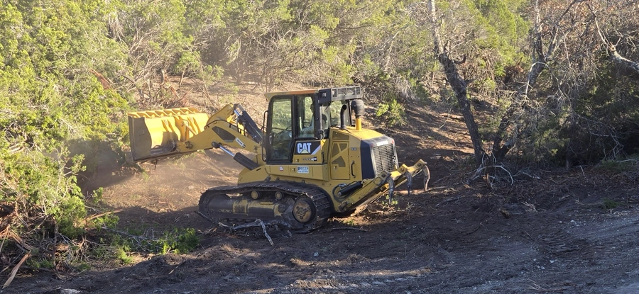 A large yellow Caterpillar bulldozer working on clearing and leveling a wooded area. The machine is moving through the underbrush, creating a path through the dense foliage and fallen branches.