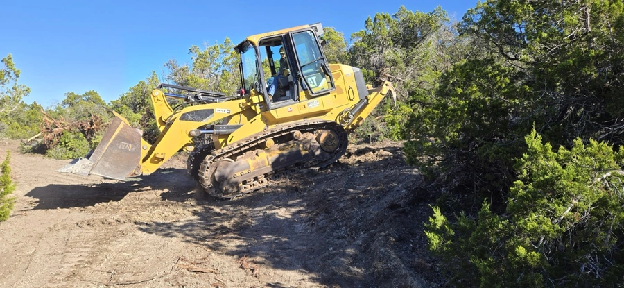 A yellow Caterpillar bulldozer tractor is seen moving earth and debris on a construction site surrounded by trees and vegetation.