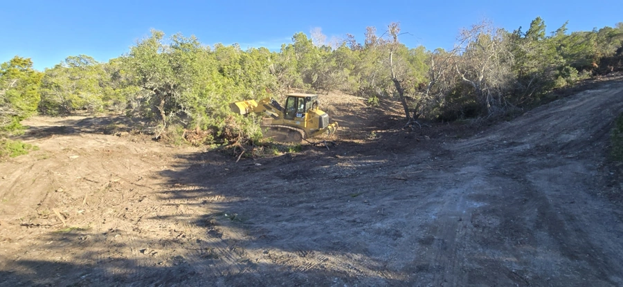 A yellow bulldozer clearing debris and vegetation from a wooded area, preparing the land for construction or development. The image shows the bulldozer working on a dirt road surrounded by trees and bushes.
