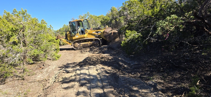 A yellow bulldozer excavating and clearing a dirt path through a wooded area with lush foliage and a blue sky overhead.