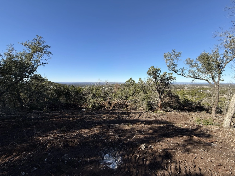 A rugged, mountainous landscape with trees and a cleared dirt path, overlooking a scenic valley in the distance under a clear blue sky.