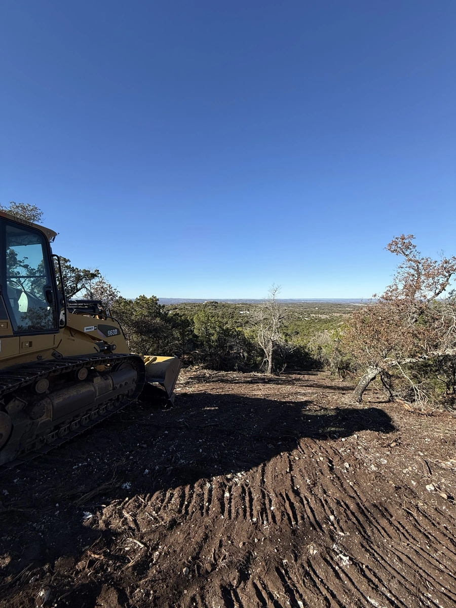 A Caterpillar tracked excavator is seen working to clear and prepare a rural, wooded site for future construction. The heavy machinery is removing vegetation and earthmoving in an elevated location with views of the surrounding countryside.