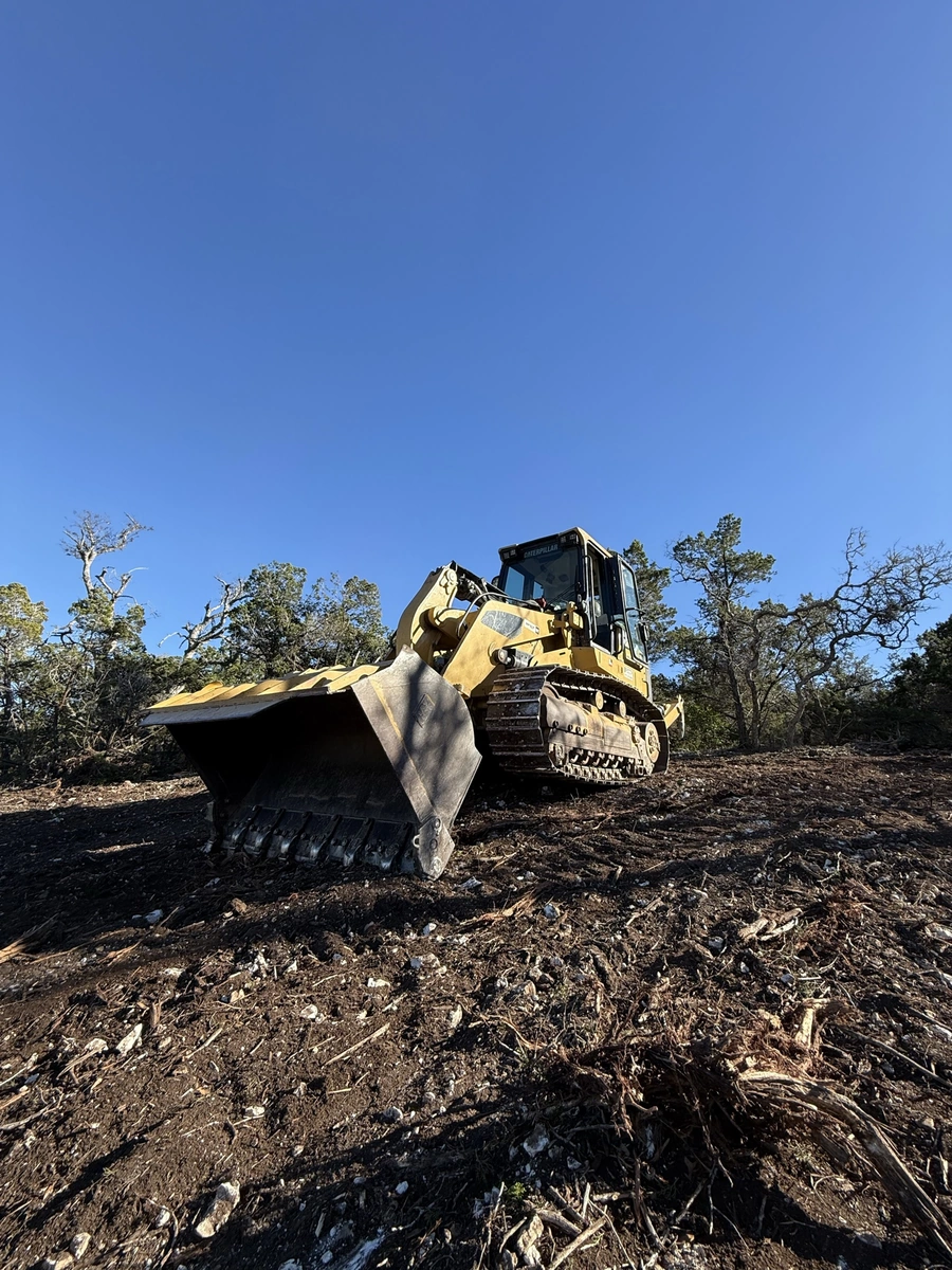 A large yellow bulldozer or track loader excavator working on clearing and grading a wooded, debris-filled site under a bright blue sky.