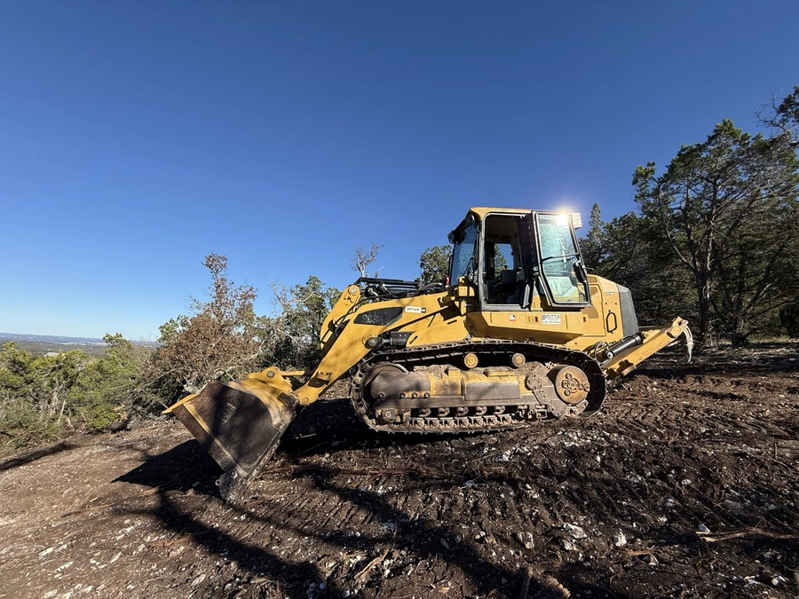 A heavy construction bulldozer with a large front loader bucket, operating in a wooded, rural area with a clear blue sky overhead. The bulldozer appears to be actively moving earth and clearing vegetation as part of a land improvement or construction project.