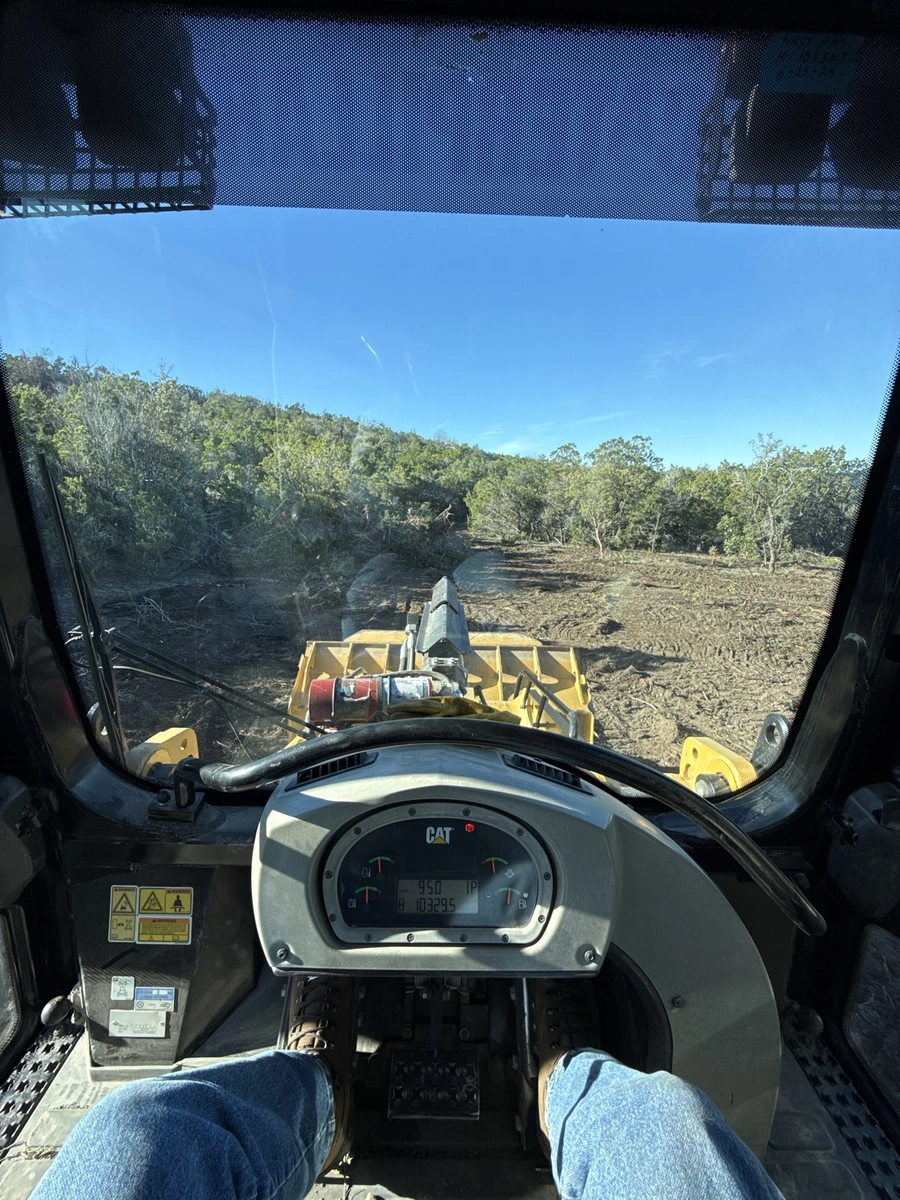 View from the operator's seat of a heavy machinery vehicle, showing a dirt construction site with trees and a clear blue sky in the background.