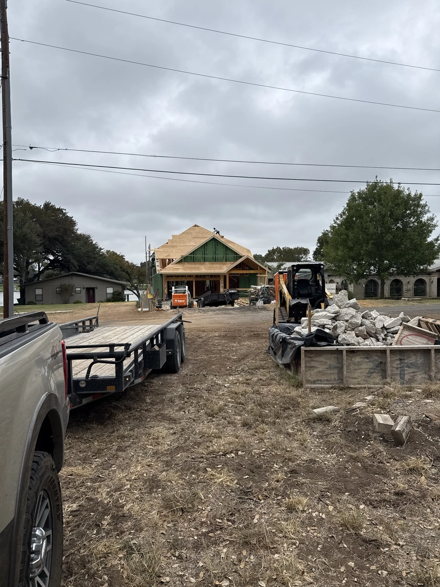 A construction site with heavy machinery, materials, and a partially built structure. The image shows an active work environment with a truck, bulldozer, and piles of rocks and other construction materials.