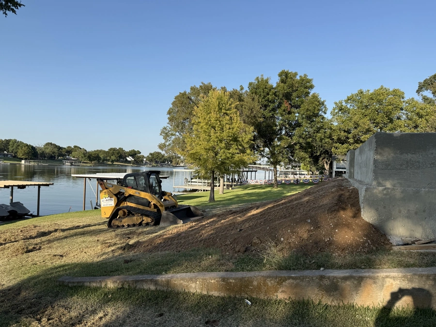 A yellow skid steer bulldozer on a dirt mound next to a lakefront property with trees and buildings in the background.