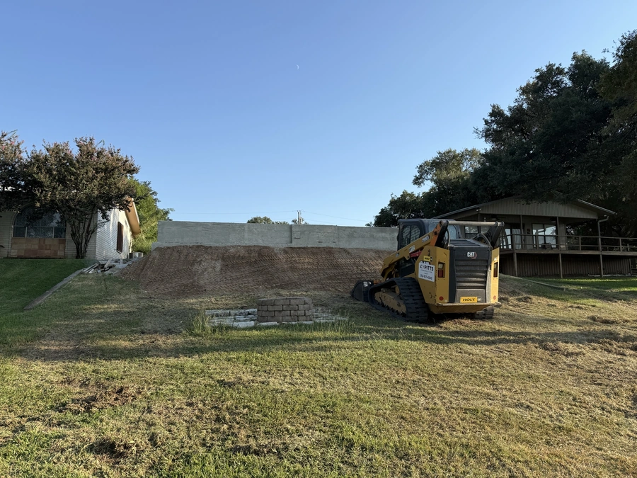 A large construction vehicle, likely a skid steer or small bulldozer, operating on a grassy area with a partially constructed retaining wall in the background. The scene suggests land clearing, site preparation, or excavation work being performed at a construction site.