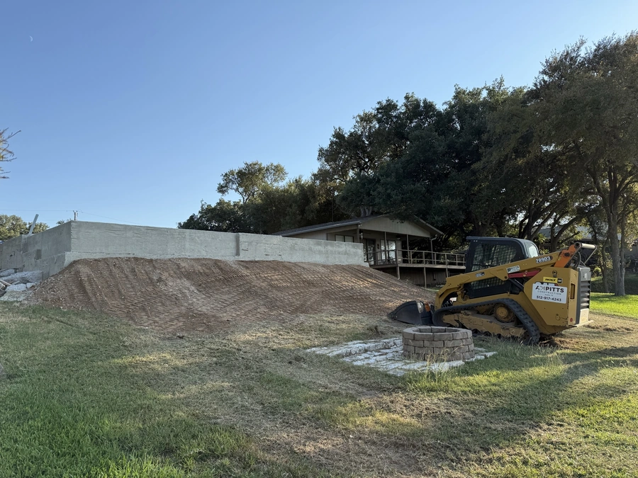 A construction site with a large yellow bulldozer in the foreground, preparing the land for a new project. In the background, there is a small building with a covered porch and trees surrounding the area.