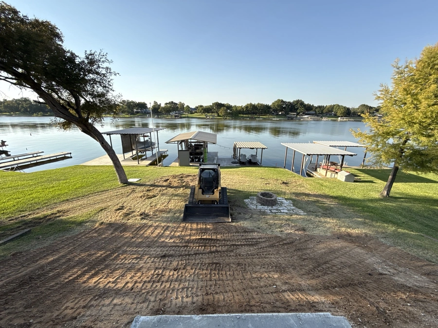 A peaceful lakeside scene with a boat dock, picnic tables, and a tree-lined shoreline. In the foreground, there is a piece of construction equipment, suggesting ongoing land improvement or site work.
