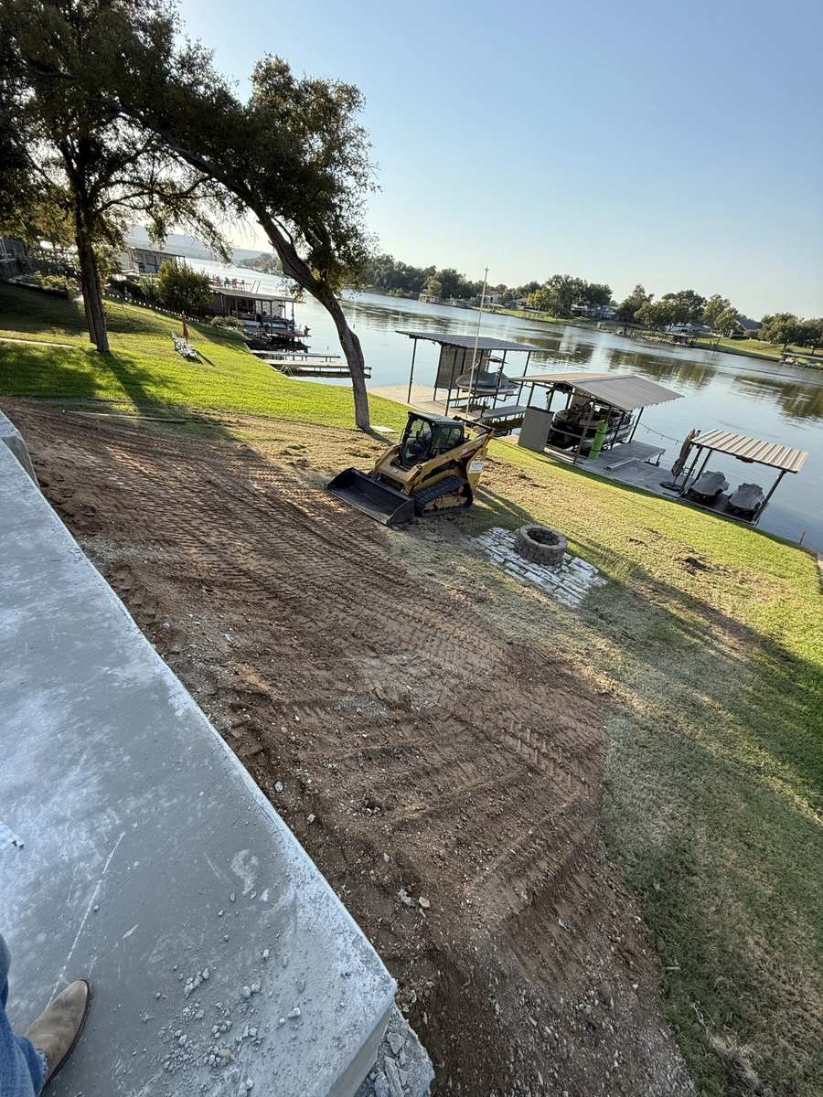 A bulldozer working on a construction site along a riverfront, with boats and buildings visible in the background. The scene shows ongoing land development and improvement work.
