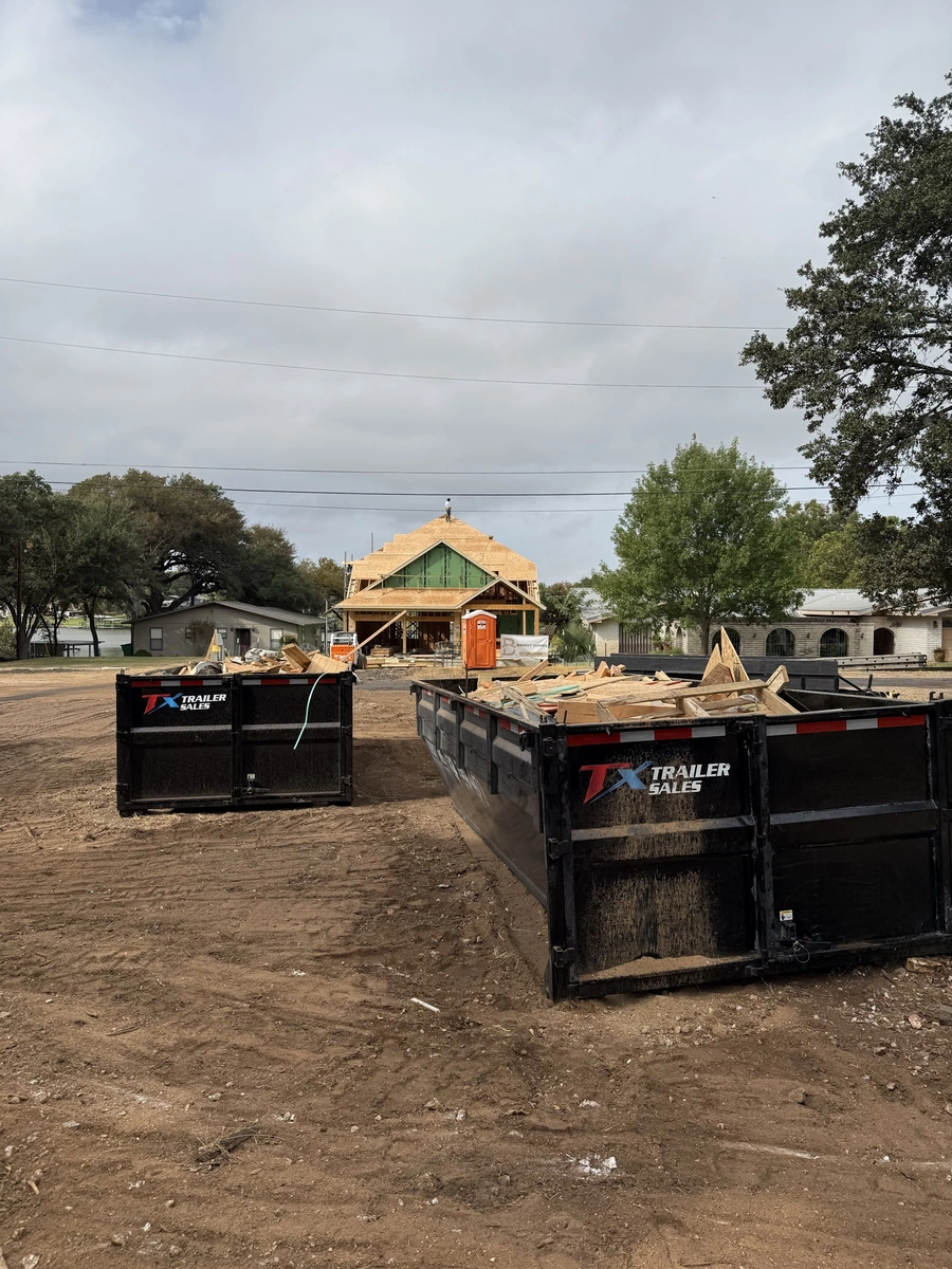 Construction site with dump trucks, trailers, and a partially built structure in a rural wooded setting, indicating site preparation work for a new development project.