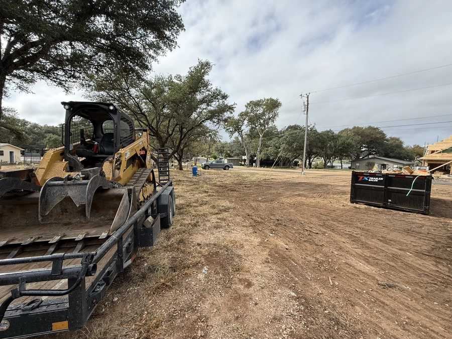 A construction site with a yellow tracked excavator or backhoe loader in the foreground, surrounded by trees and other construction equipment like a trailer. The site appears to be a clearing or preparation for a new development or project.