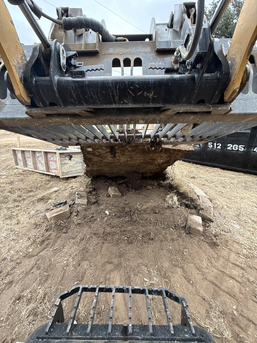 Image shows the underside of a heavy construction vehicle, likely an excavator or backhoe, digging into the ground. The machine's treads and various hydraulic arms and attachments are visible, covered in dirt and debris from the work being performed. This appears to be a construction site, with the vehicle actively engaged in earthmoving or site preparation activities.