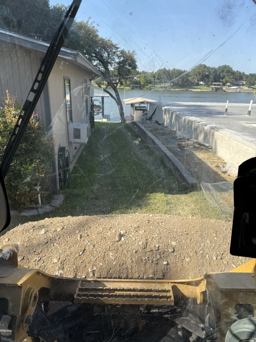 Image showing a construction site with a small building and a lake or waterway in the background. A heavy equipment machine is visible in the foreground, with dirt and debris on the ground.
