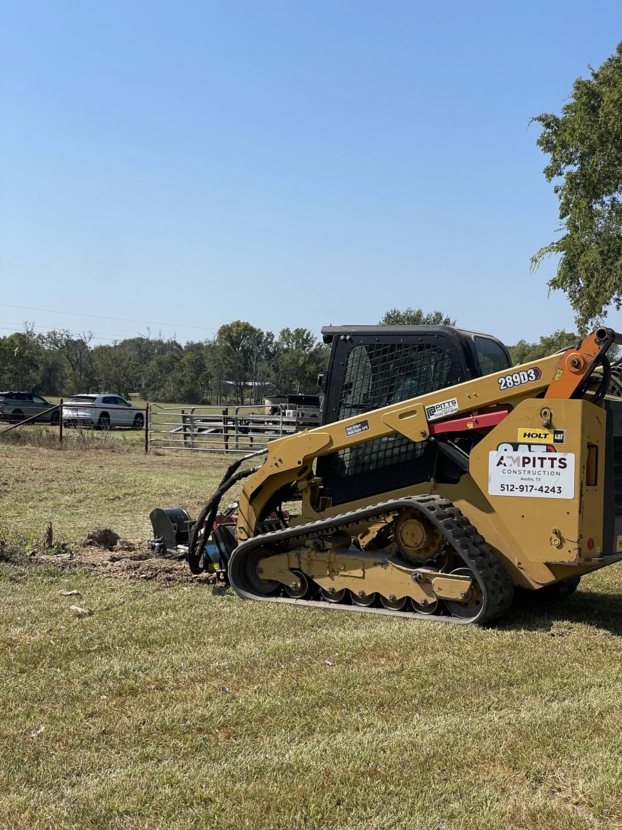 A yellow skid steer loader with the A.M. Pitts Construction logo on it, working on a grassy field with trees in the background. A pickup truck is visible in the distance.