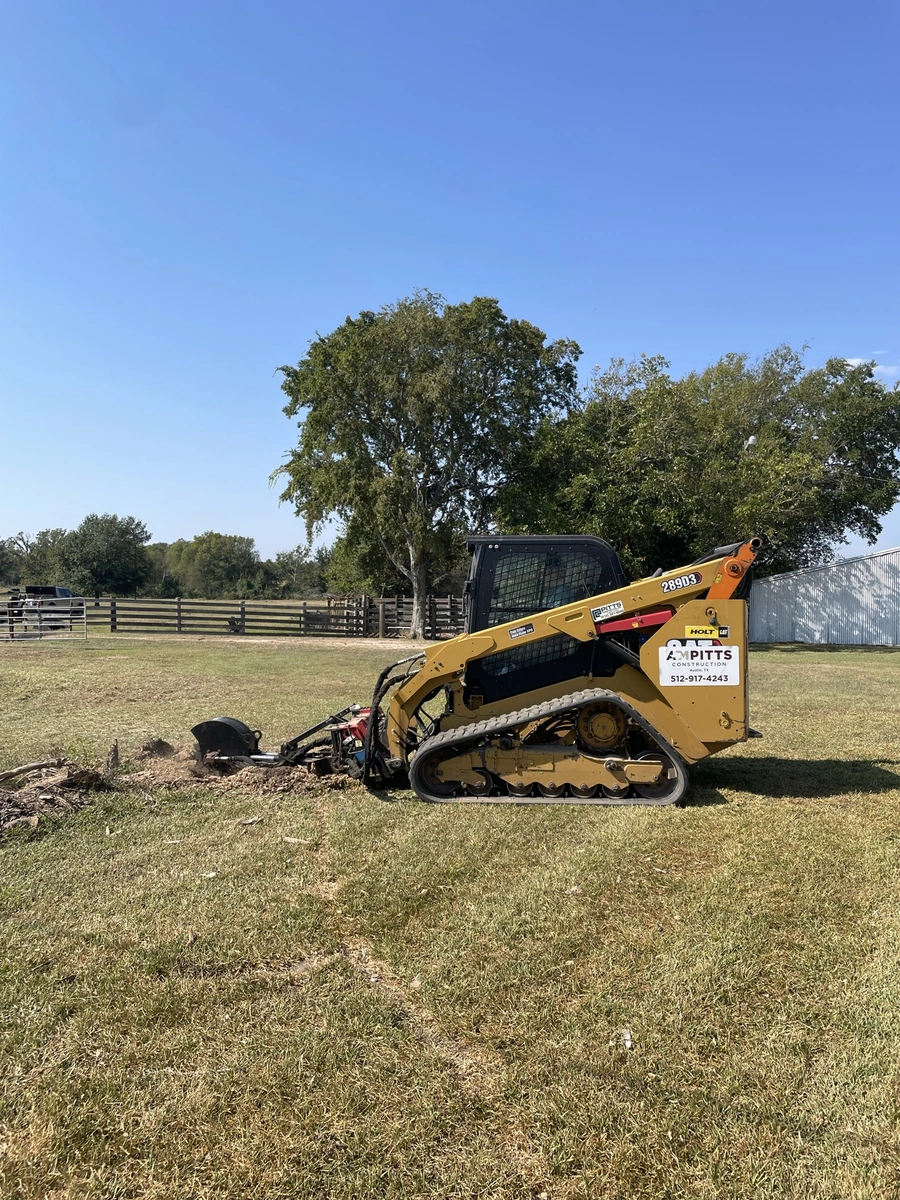A large yellow tracked skid steer loader used for land clearing and site preparation work on a grassy field surrounded by trees and fencing.