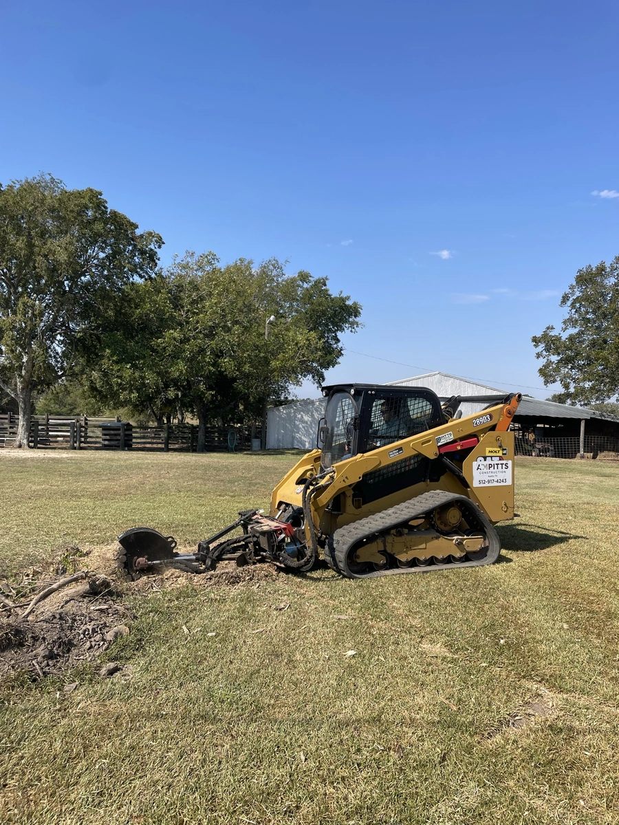 A yellow skid steer loader with a rear-mounted attachment working on a grassy field, surrounded by trees and a barn-like structure in the background.