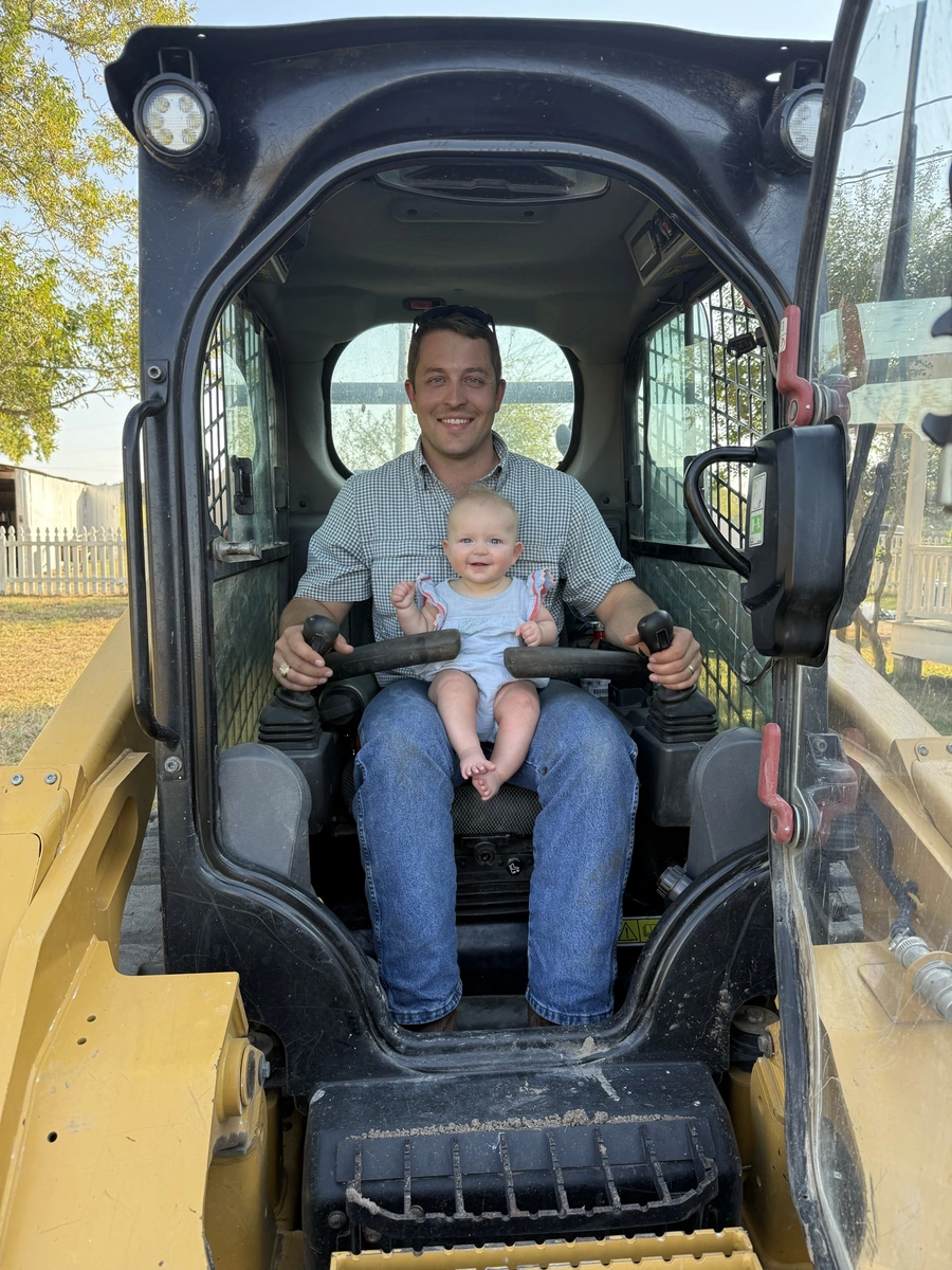 A smiling man holding a happy baby while sitting in the driver's seat of a large construction vehicle or tractor.