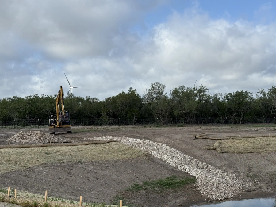 An excavator working on a construction site surrounded by piles of gravel and debris, with wind turbines visible in the background against a cloudy sky.