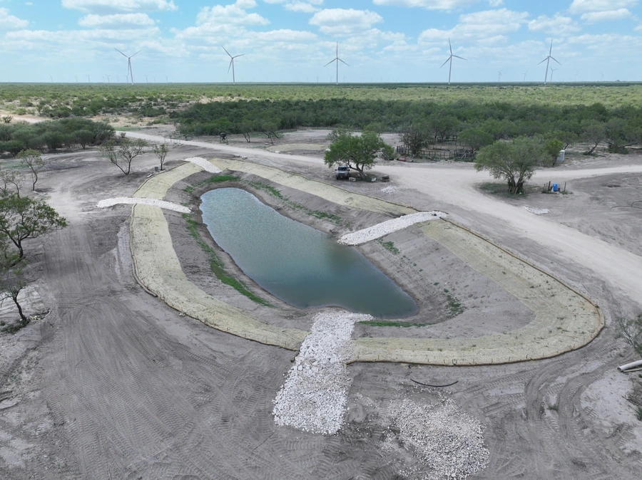 Aerial view of a large, arid landscape with wind turbines in the background and a man-made pond or reservoir in the foreground, surrounded by dirt and gravel work areas.
