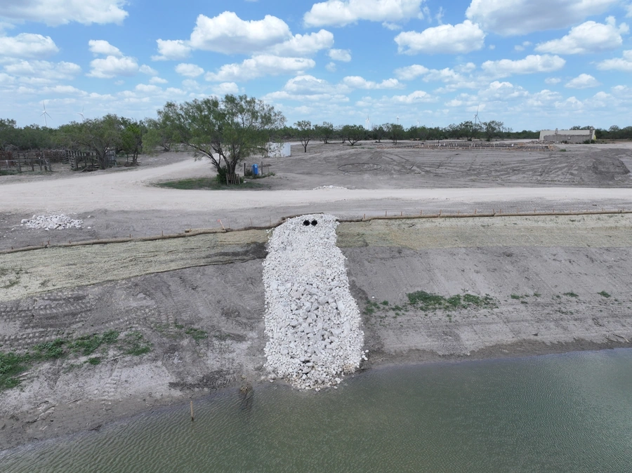 An expansive construction site with a dirt road, gravel piles, and machinery in the background. A large mound of crushed limestone or similar material is visible in the foreground.