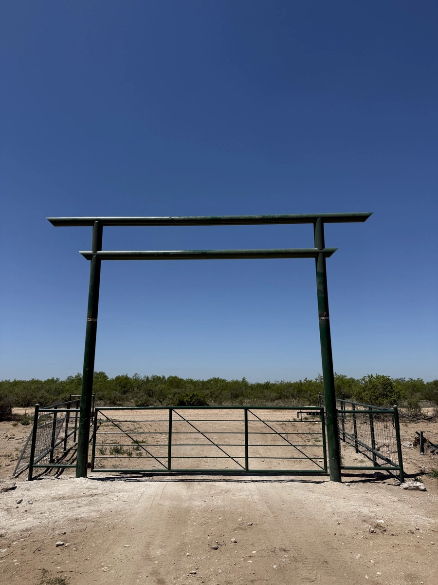 A large metal gate frame in a rural, desert-like landscape with some vegetation in the background.