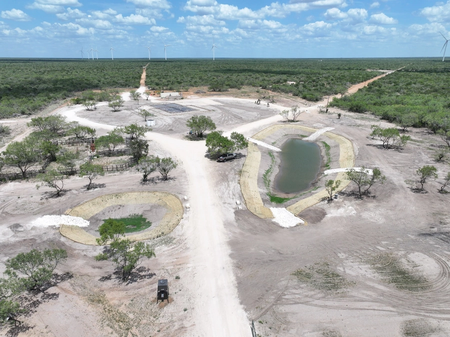 Aerial view of a large construction site in a remote, rural area with wind turbines visible in the background. The site features multiple earthworks, ponds, and staging areas for heavy equipment.