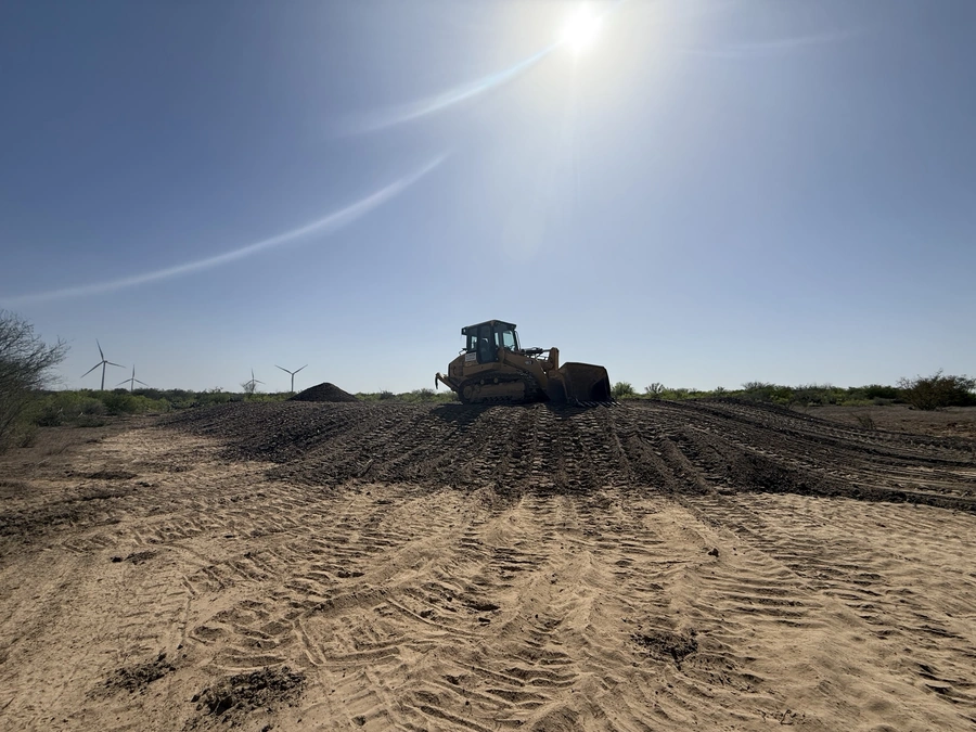 A large bulldozer tractor moving soil and preparing a construction site under a bright, sunny sky with wind turbines visible in the background.