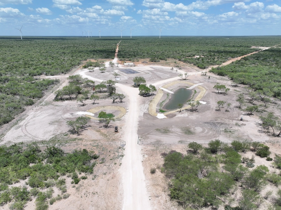 An aerial view of a construction site in a rural, remote area. The site features large cleared areas, dirt roads, and several freshwater ponds or reservoirs. Wind turbines can be seen in the background, indicating the use of renewable energy sources. This image showcases the company's expertise in land improvement, site work, and excavation projects in remote, natural environments.