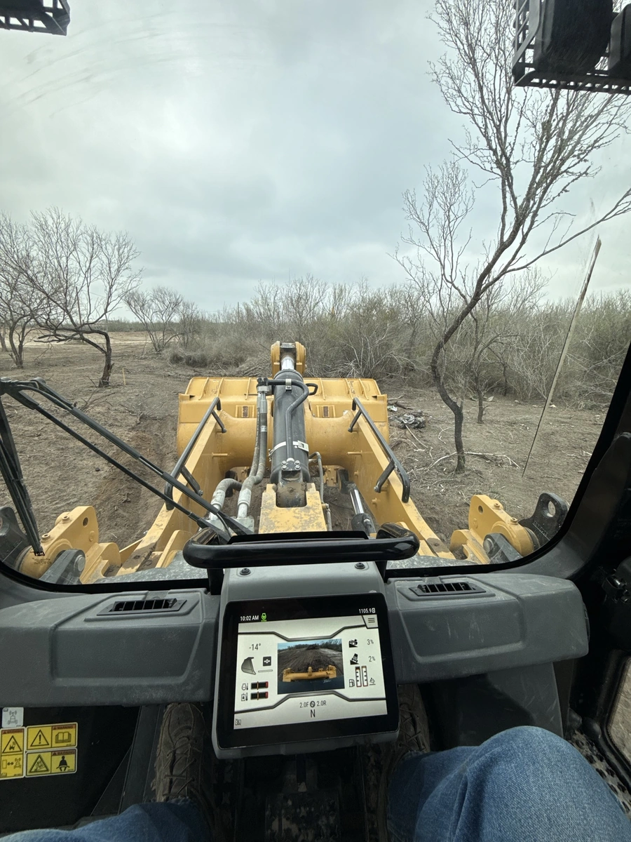 View from the cab of a large yellow excavator machine on a remote, wooded site. The excavator's bucket and hydraulic arms are visible in the foreground, with bare trees and a cloudy sky in the background.