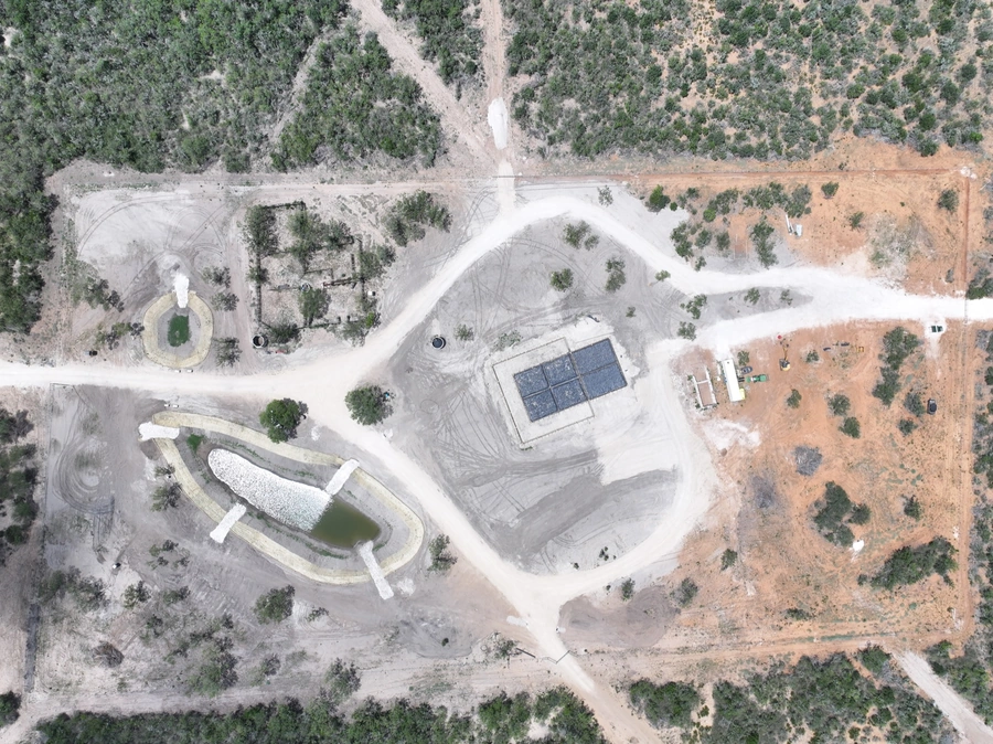 Aerial view of a construction site with a large building, earthmoving equipment, and other infrastructure scattered across a rural, forested landscape.