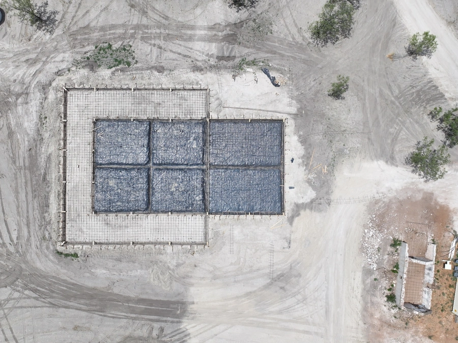 Aerial view of a construction site with a large, rectangular enclosure made of metal fencing and tarps, surrounded by a rugged, mountainous landscape with sparse vegetation.