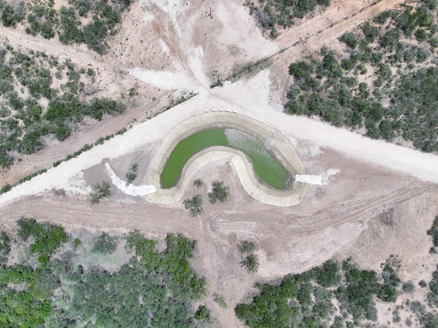 Aerial view of a completed dam construction project in a remote, wooded area. The dam has created a small, green pond surrounded by a curved, earthen embankment structure.