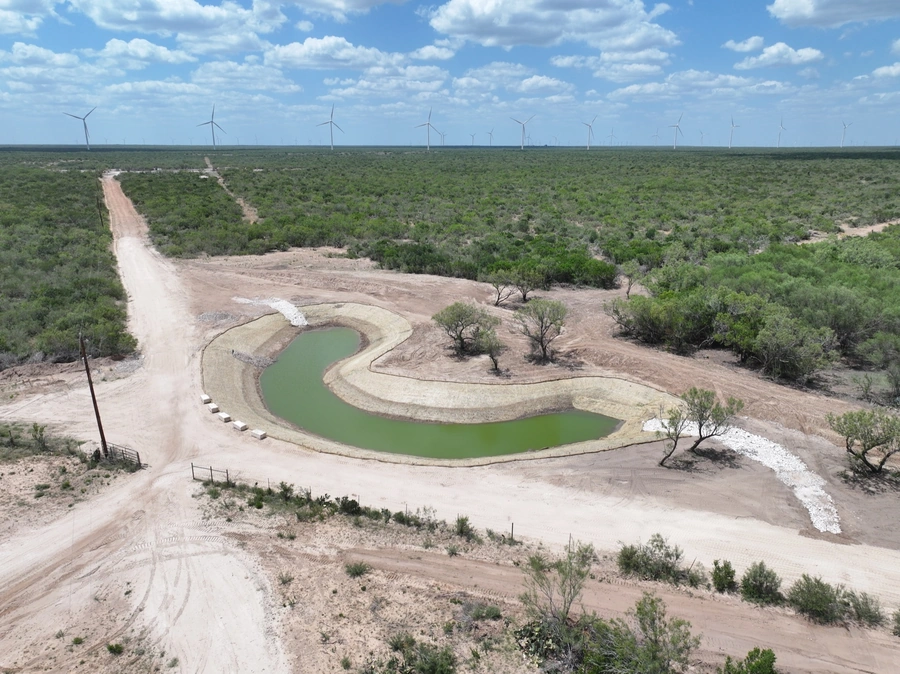Aerial view of a large wind farm surrounded by a green, lush landscape with a winding dirt road and a pond in the foreground. The scene depicts renewable energy production in a natural environment.