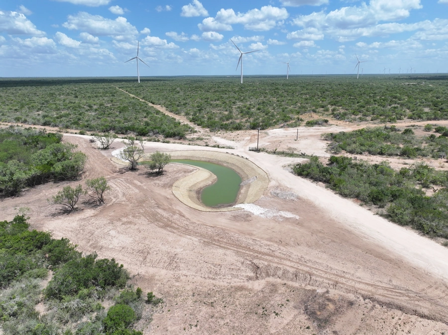 Aerial view of a large wind farm in a dry, arid landscape with a curved dirt road and small pond or watering hole in the foreground.