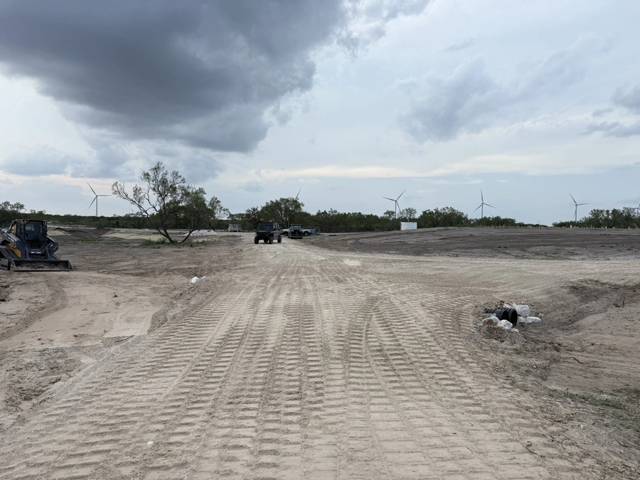 An open construction site with heavy machinery, wind turbines in the background, and a dirt road leading through the landscape under a cloudy sky.