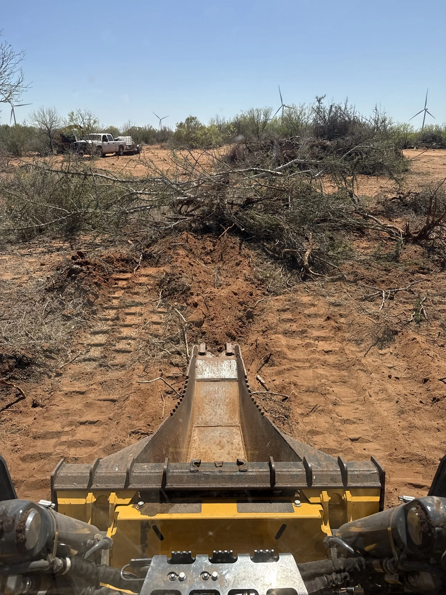 An image showing a construction site with a bulldozer in the foreground, clearing debris and vegetation. In the background, several wind turbines are visible, indicating the location is likely a rural, remote area.