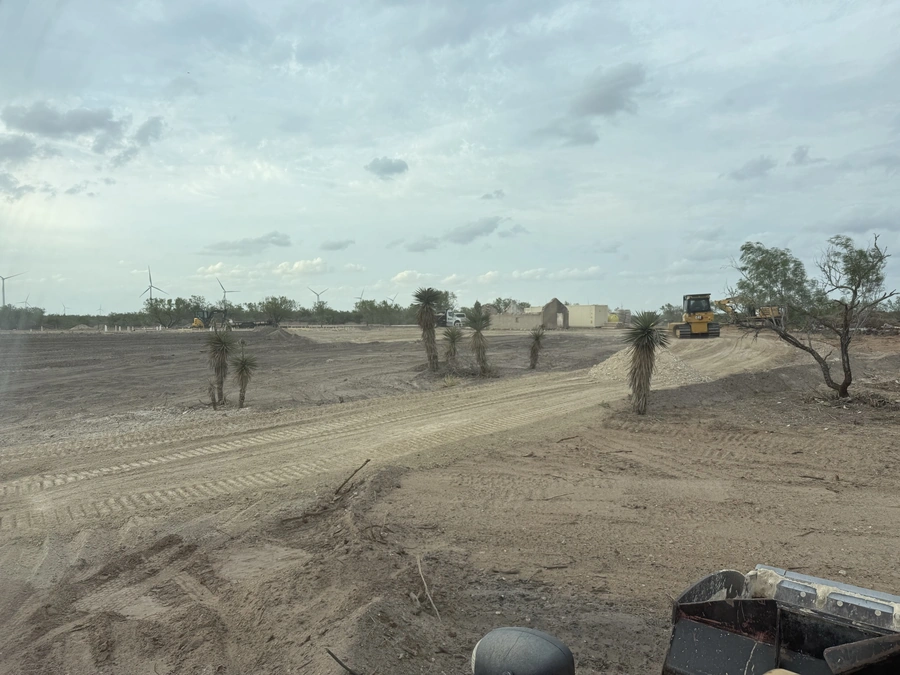 An expansive, arid landscape with scattered palm trees and a construction site in the distance. Heavy machinery and equipment can be seen moving through the dusty terrain.