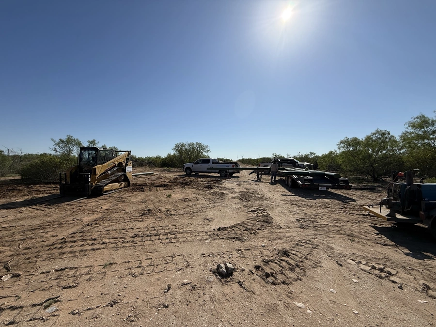 A construction site with a bulldozer, trucks, and workers visible in a rural, wooded setting under a bright, blue sky.