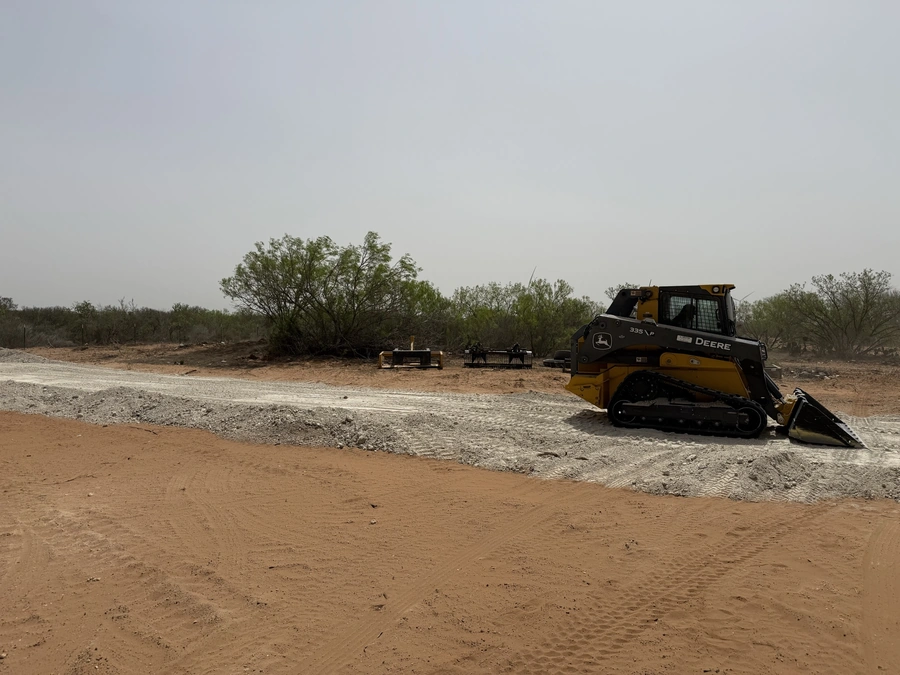 A John Deere skid steer loader clearing and grading a dirt road through a wooded area on a construction site.