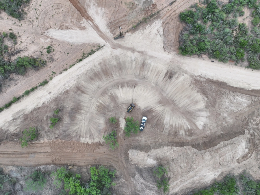 Aerial view of a construction site with heavy machinery and earthworks in progress. Large areas of disturbed soil and earthen berms can be seen, surrounded by lush greenery and natural vegetation.