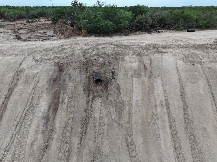 An aerial view of a remote, rural area with a dirt road and thick vegetation surrounding it. The road shows signs of heavy machinery and construction work, with tire tracks and uneven terrain visible.