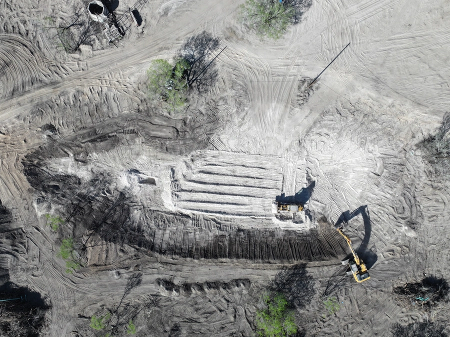 Aerial view of an active construction site with heavy machinery and earthmoving equipment working on clearing and grading the land. The image shows the dramatic scale and scope of the site work being performed.