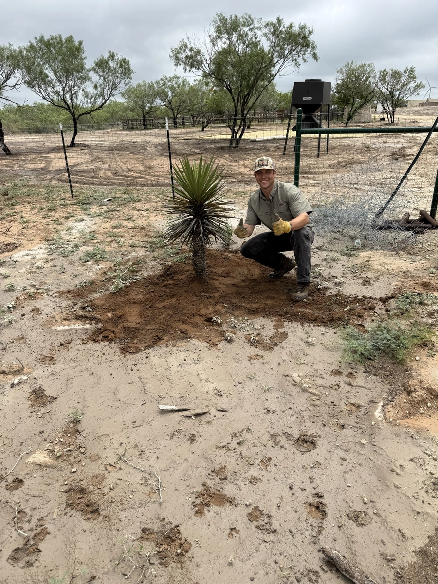 A construction worker, wearing a hat and gloves, is digging in the dirt at a rural site with trees and a fence in the background.